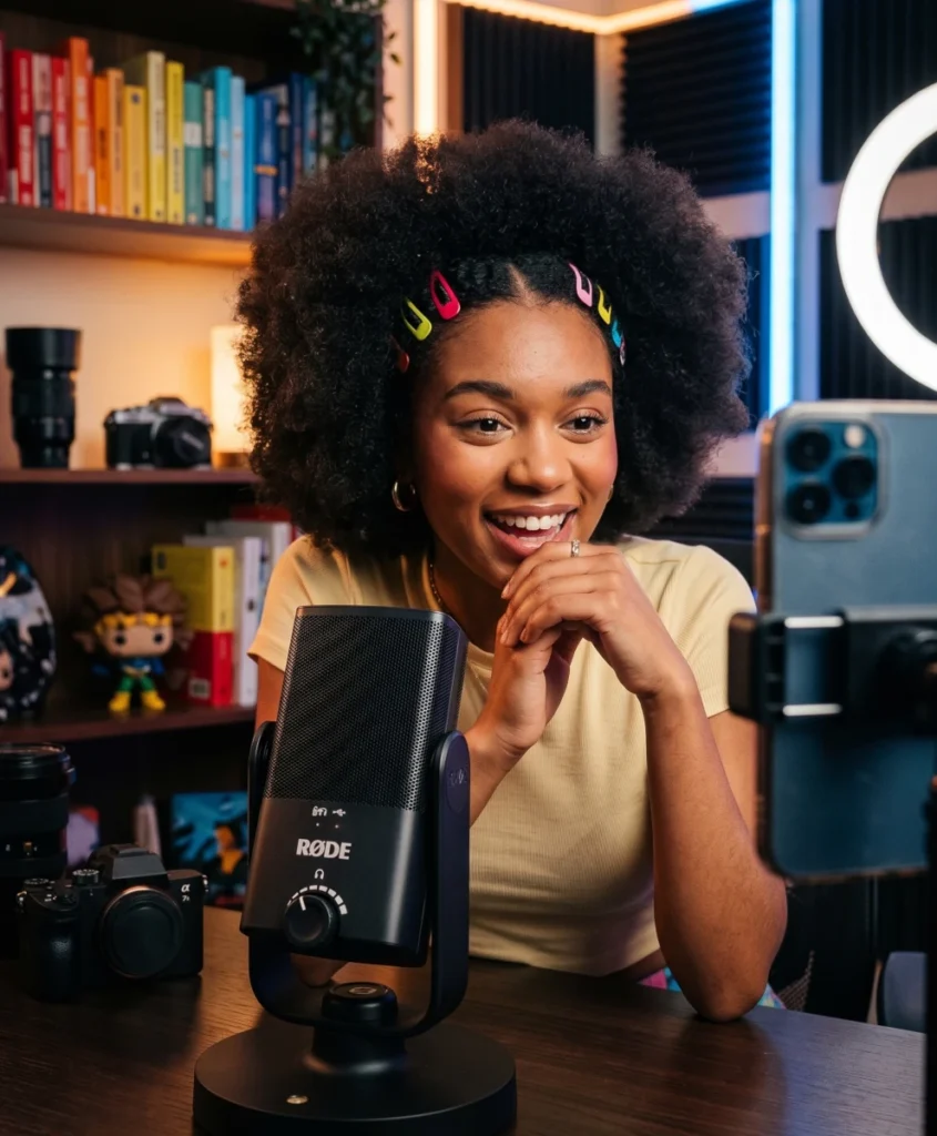 Young Black female content creator recording a video with a RØDE NT-USB Mini microphone on her desk and a smartphone on a tripod