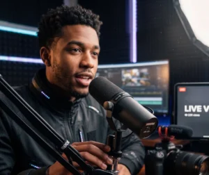Young man speaking into a Shure SM7B dynamic microphone mounted on a boom arm in a studio setup with acoustic foam walls and video editing monitors.